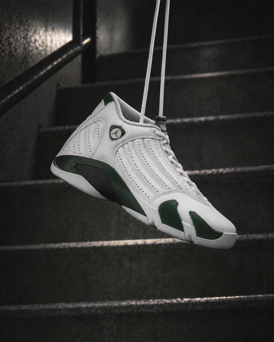 Air Jordan 14 Forest Green white and green sneaker suspended by white laces against dark stairs